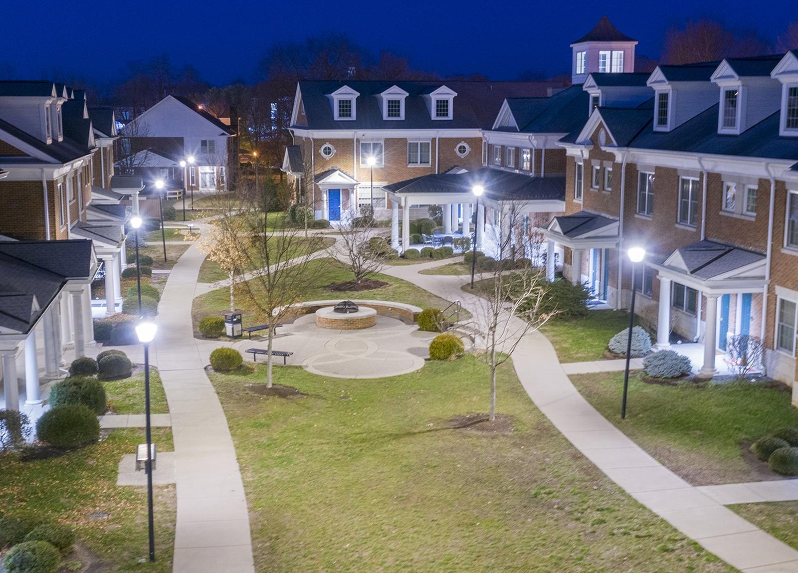 Aerial view of a well-lit courtyard at night, surrounded by brick buildings with white columns and dormer windows, featuring paths and a central round plaza.
