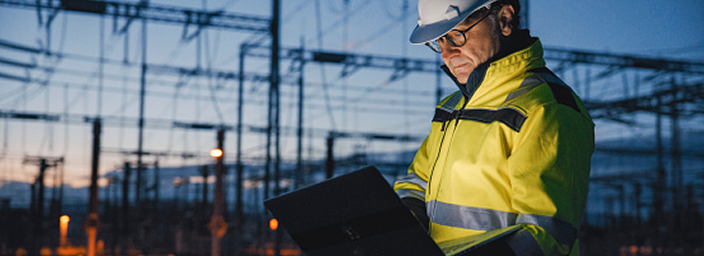 Engineer in a high-visibility jacket and helmet works on a laptop at an outdoor electrical substation during twilight.