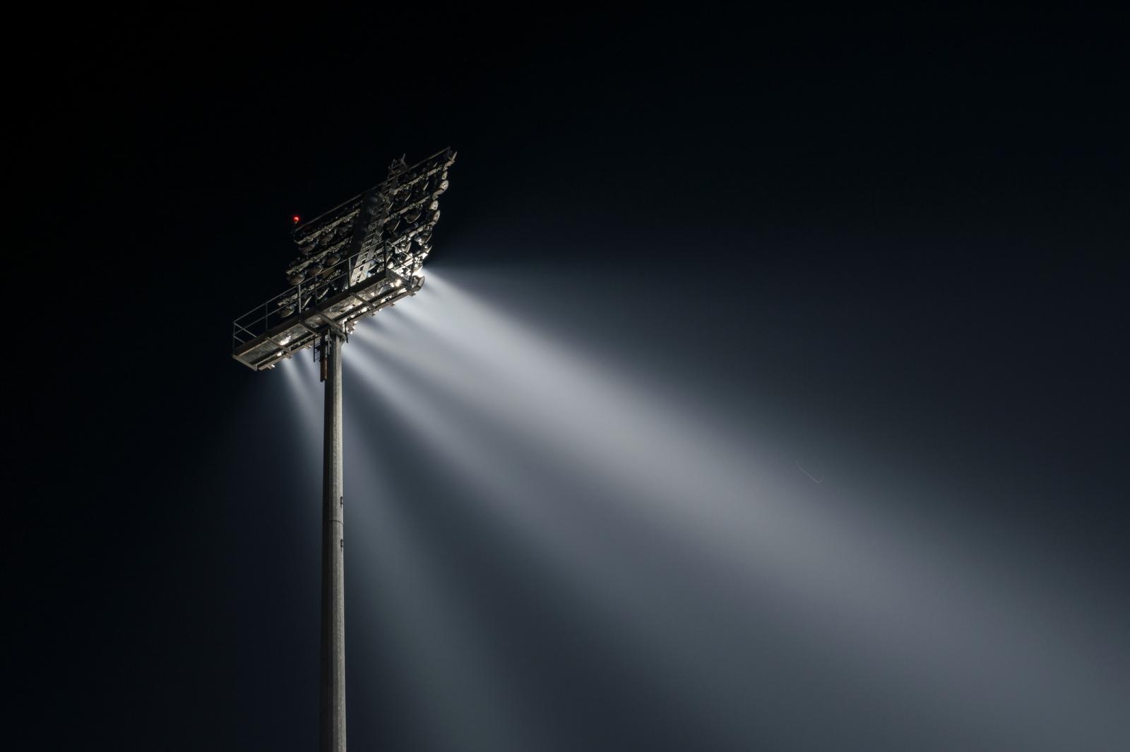 A tall floodlight tower illuminates the night sky with bright, focused beams, creating a stark contrast against the dark background.