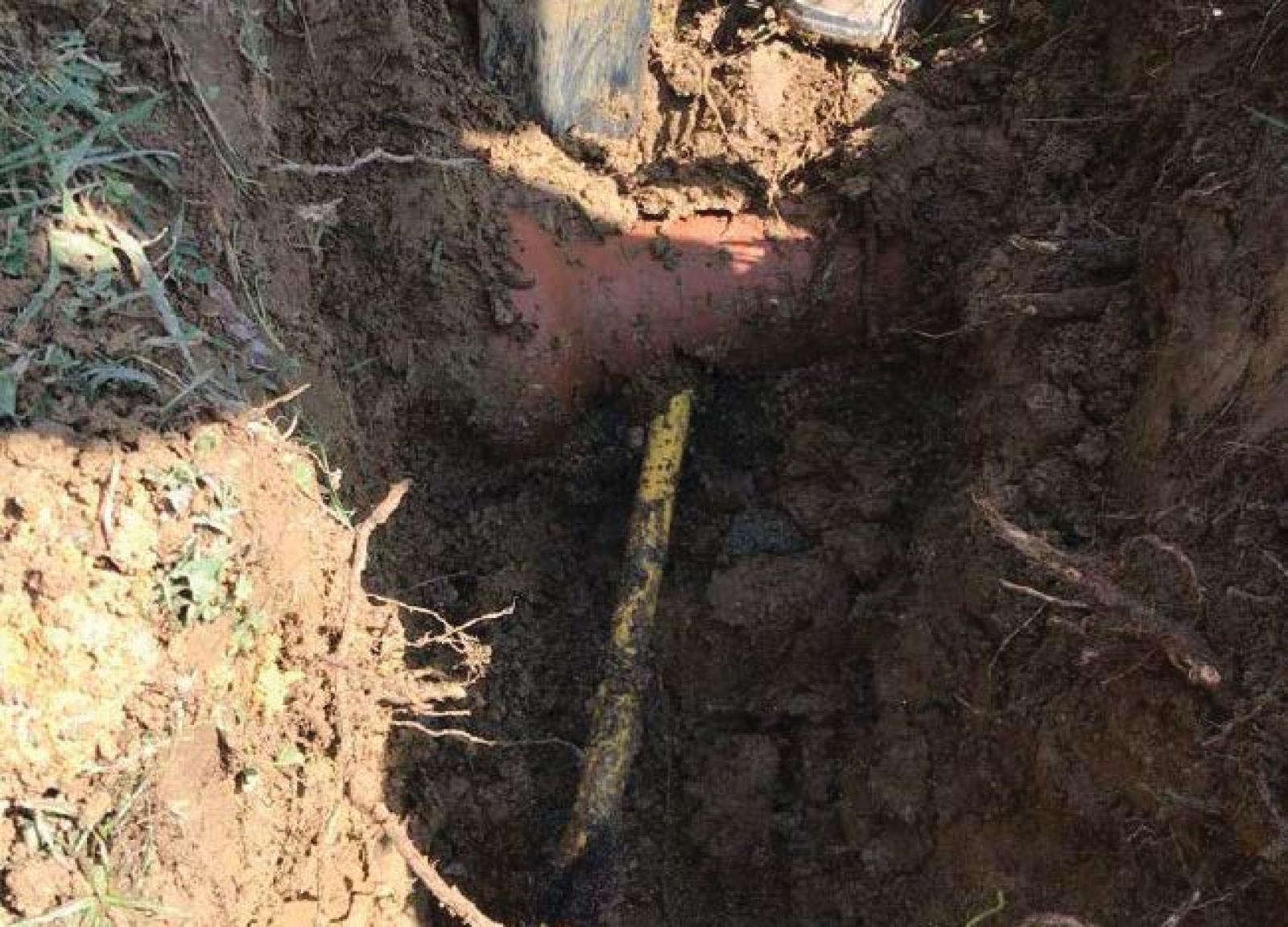 A partially buried orange pipe intersecting with a narrower yellow pipe in a dirt trench.