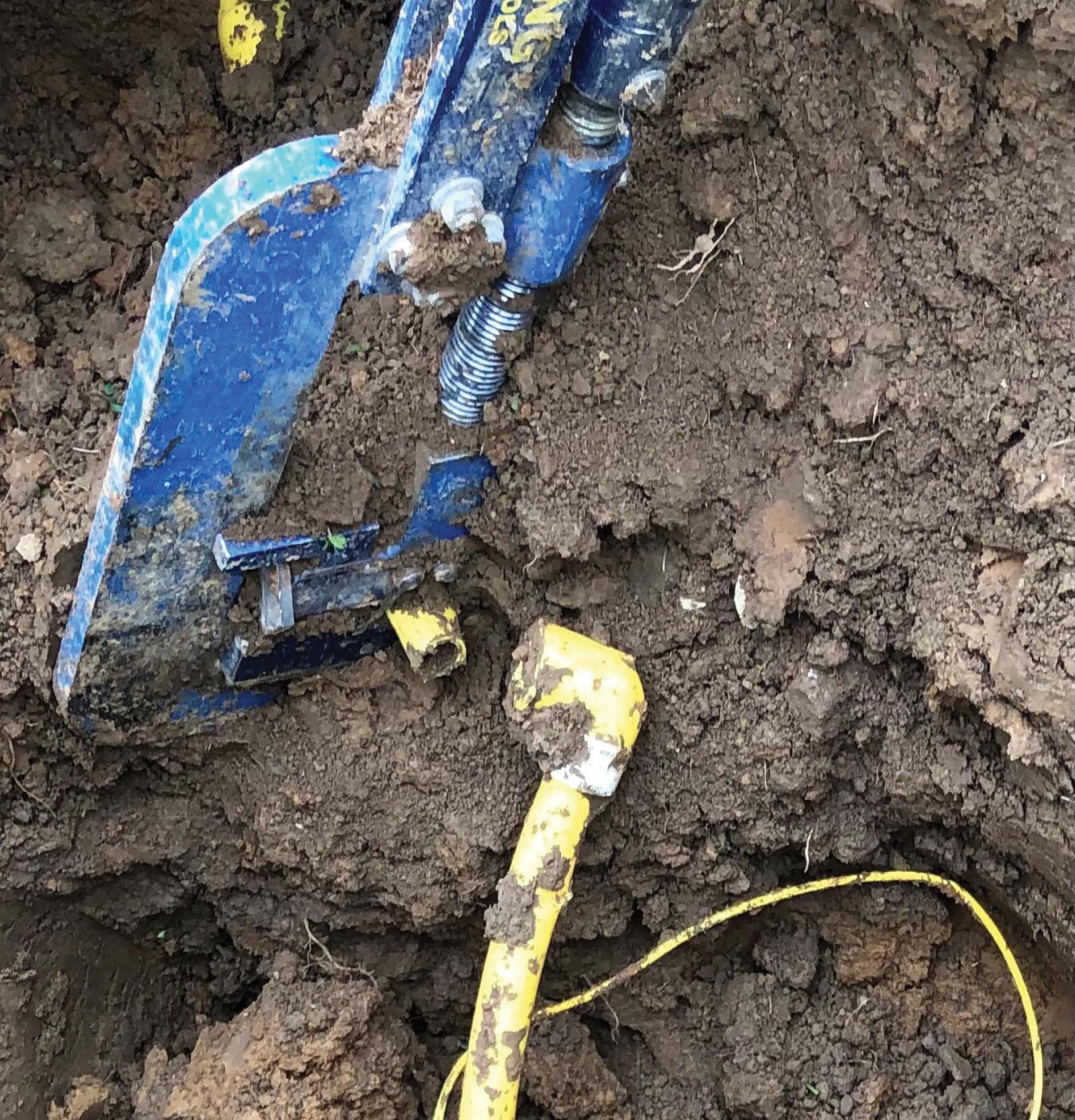 Close-up of a broken yellow pipe and blue metal tool buried in soil, depicting a broken cross-bore.