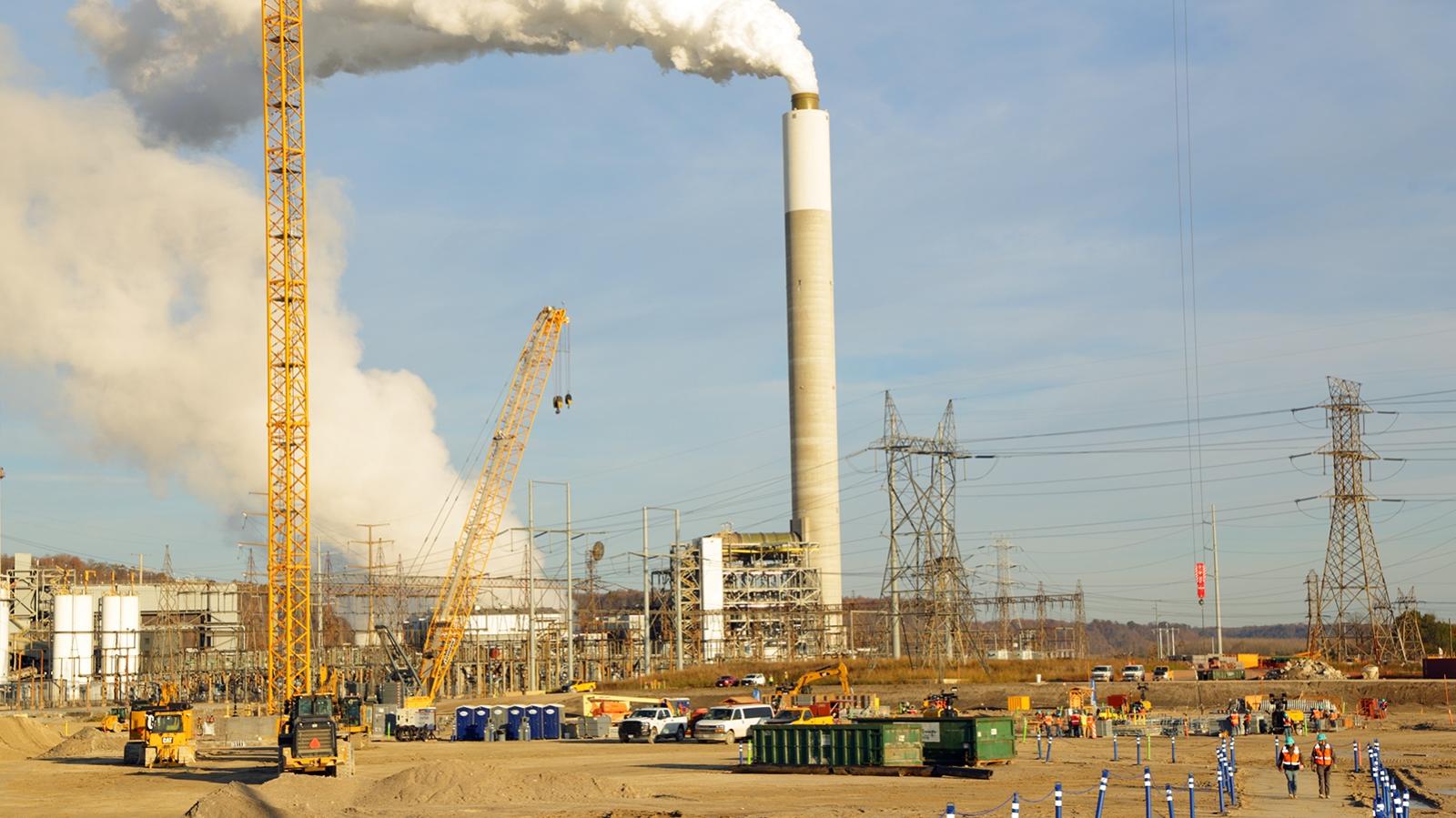 Industrial construction site with cranes, power lines, and a chimney emitting white smoke against a blue sky.
