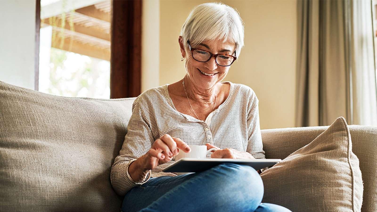 Elderly woman with glasses sits on a couch, smiling while using a tablet.