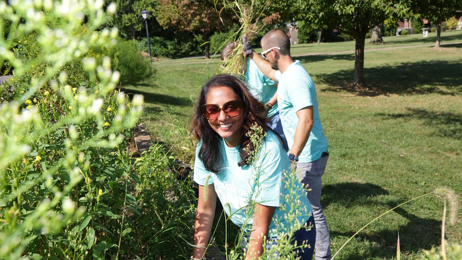 Smiling woman in sunglasses gardening with others in a park, wearing light blue T-shirts on a sunny day.