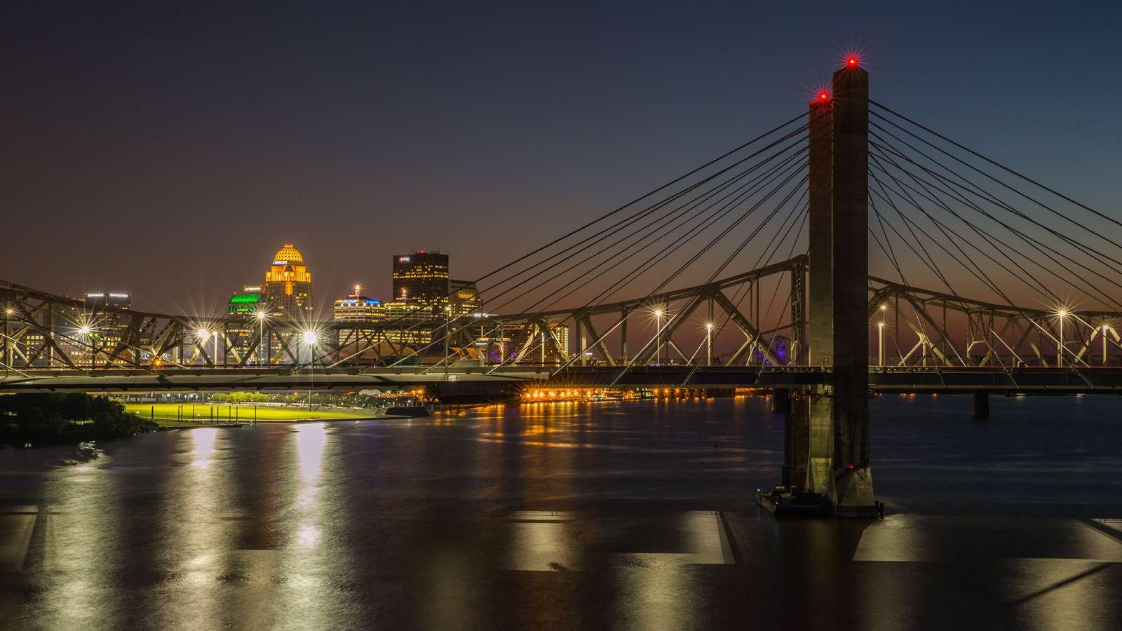 Night view of a lit cable-stayed bridge over a river with city skyline in the background.