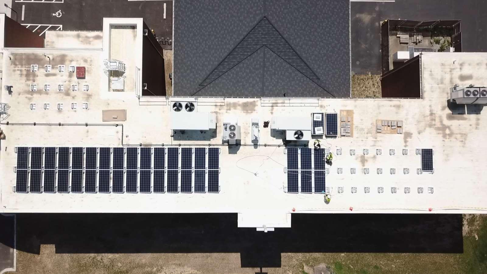 Aerial view of a rooftop with solar panels and HVAC systems on a flat white roof. Surrounding areas include parking spaces and greenery.