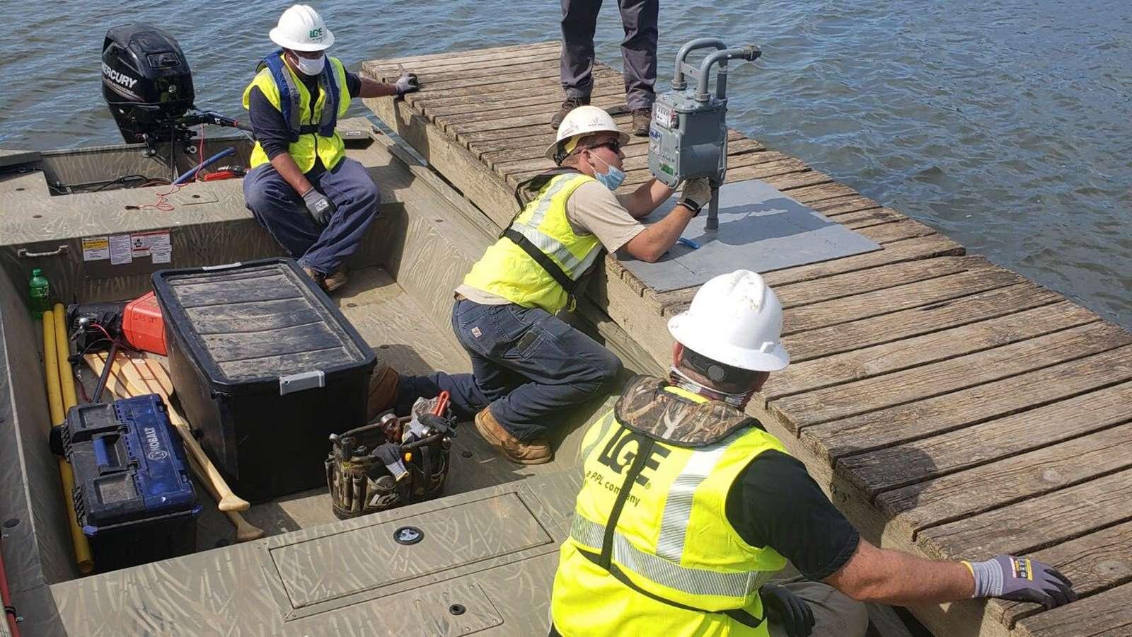 Workers in safety gear install equipment on a wooden dock by the water, with tools and storage boxes nearby.