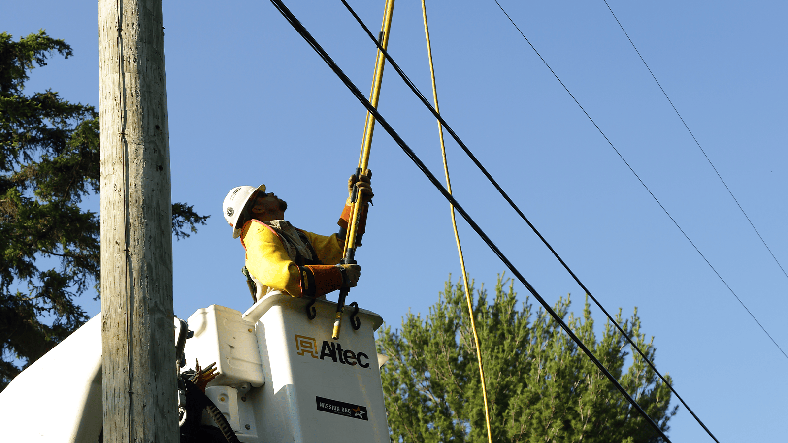 A lineman in a bucket truck repairs power lines against a clear blue sky, wearing a hard hat and safety gear.