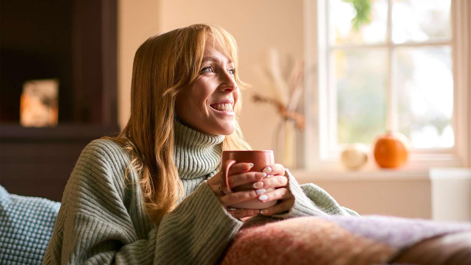 A blonde woman wearing a knit sweater, smiling and holding a mug, sits on a couch.