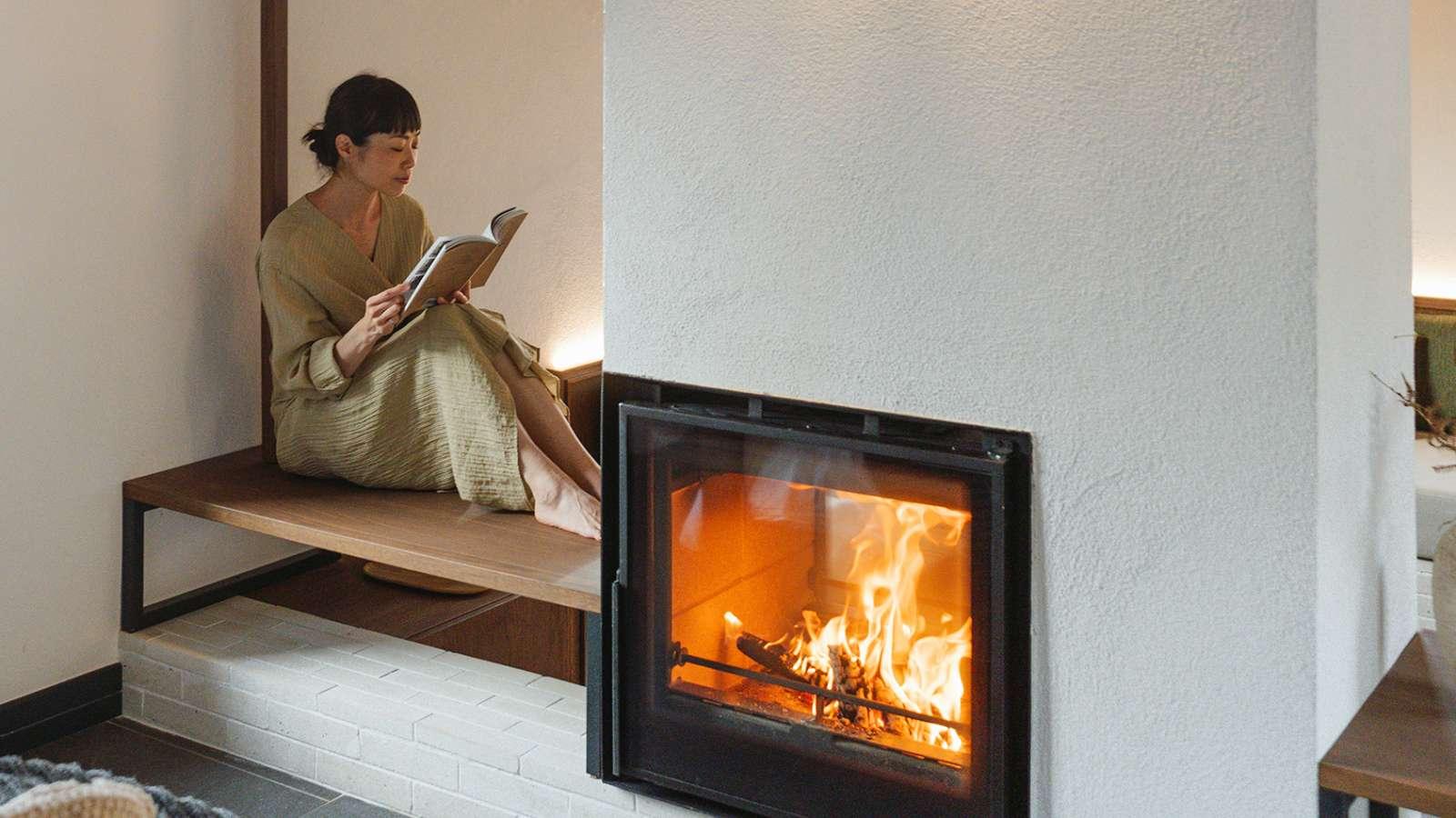 A woman reads a book while sitting on a bench next to a lit fireplace.