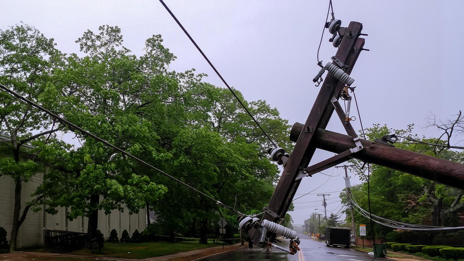 A leaning utility pole with tangled wires on a street, surrounded by greenery, under an overcast sky.