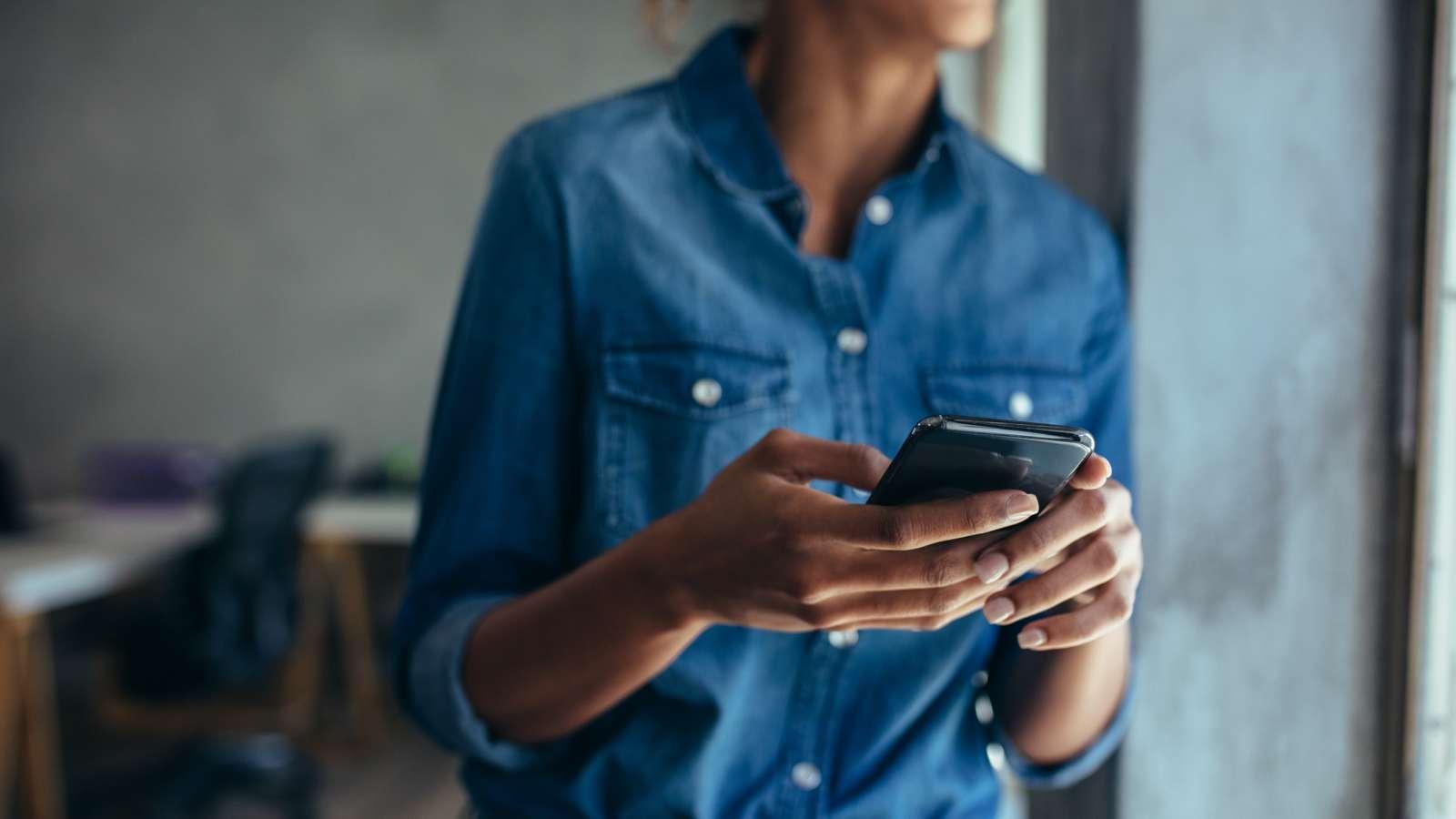Person in a denim shirt holding a smartphone and looking outside by a window, with a blurred office background.