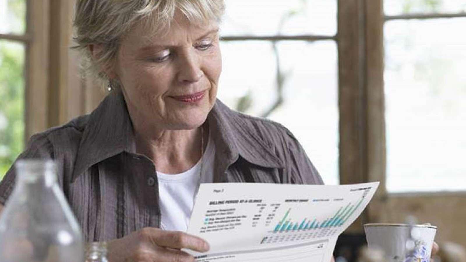 Elderly woman reading a printed graph at a table, with a blurred window background.