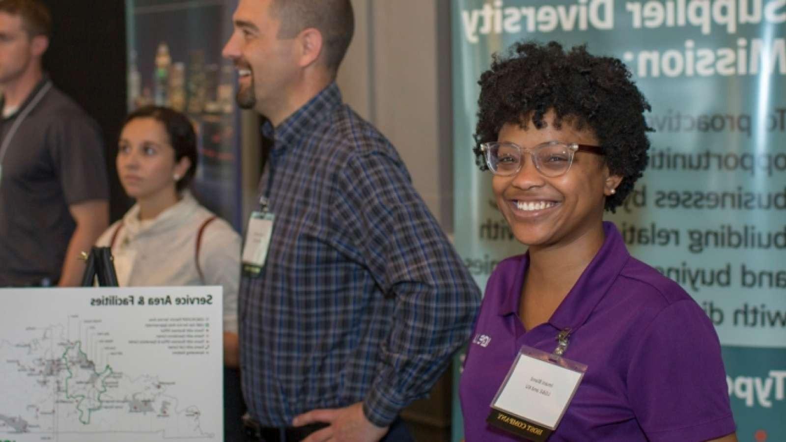 Smiling woman in a purple shirt stands near a display with a map. Three other people and a Supplier Diversity Mission sign are in the background.