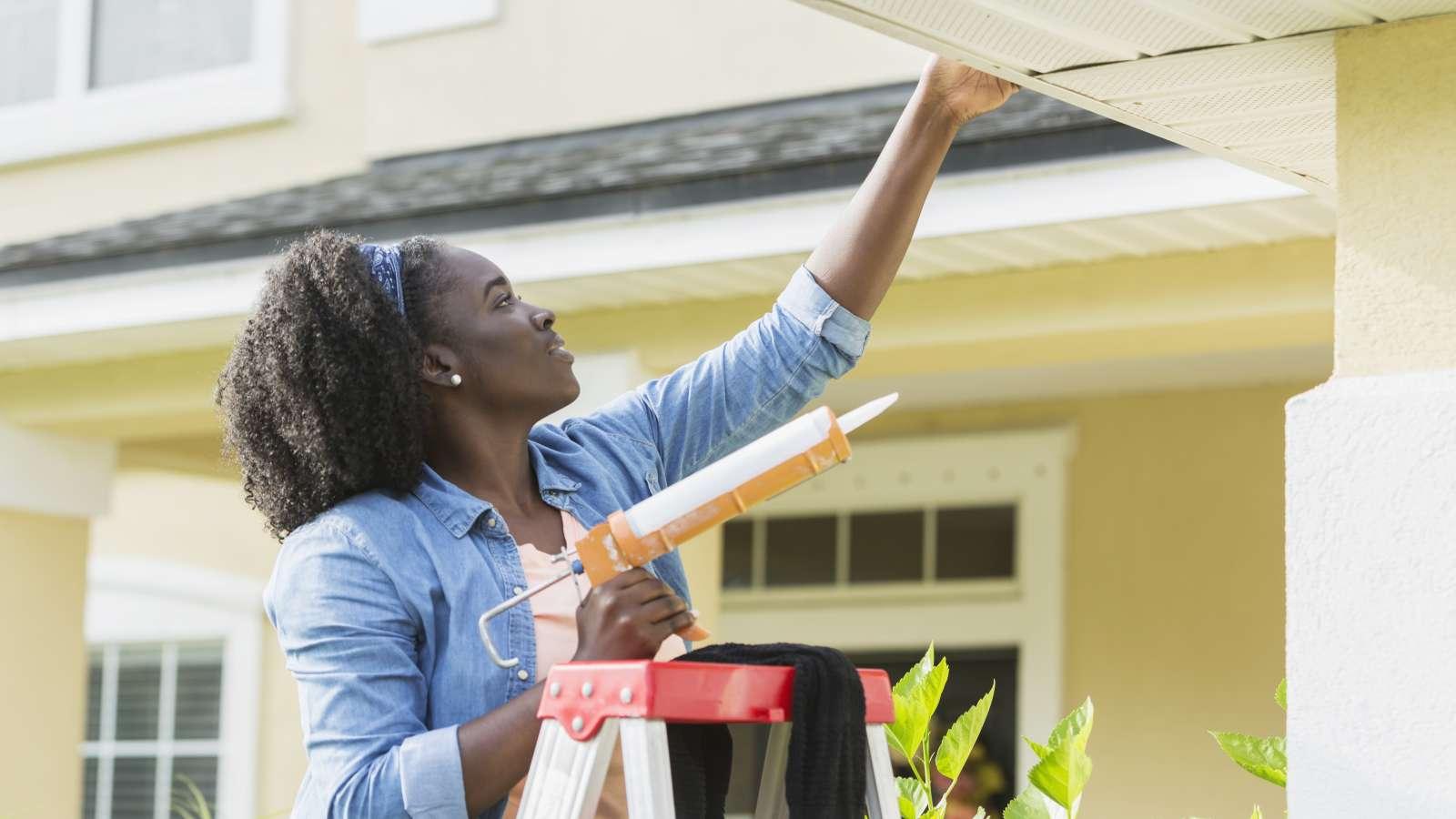 Person standing on a ladder holding a caulk gun while repairing gutters.