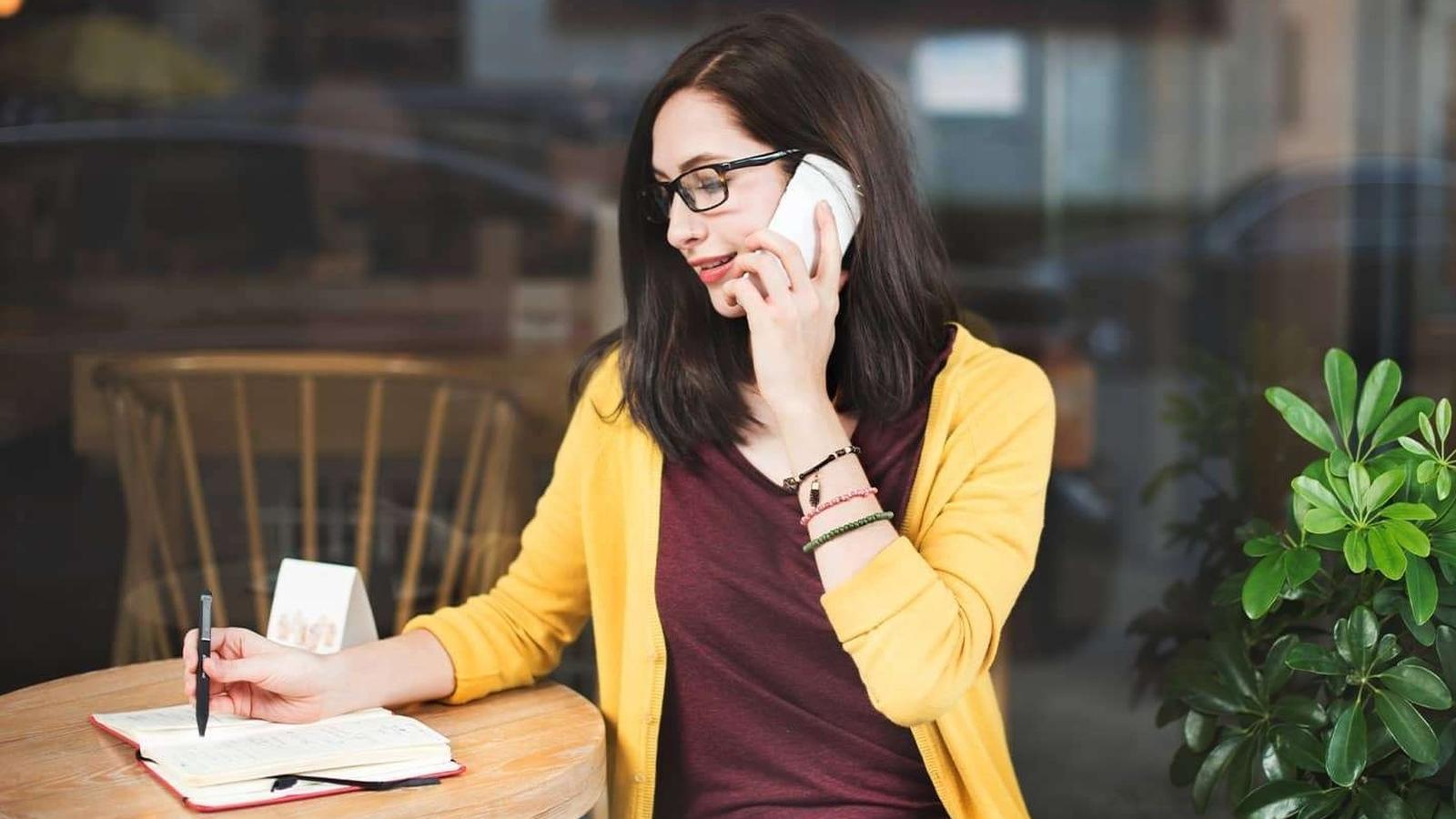 Person talks on the phone while writing in a notebook at a cafe table, with a plant and wooden chairs nearby.