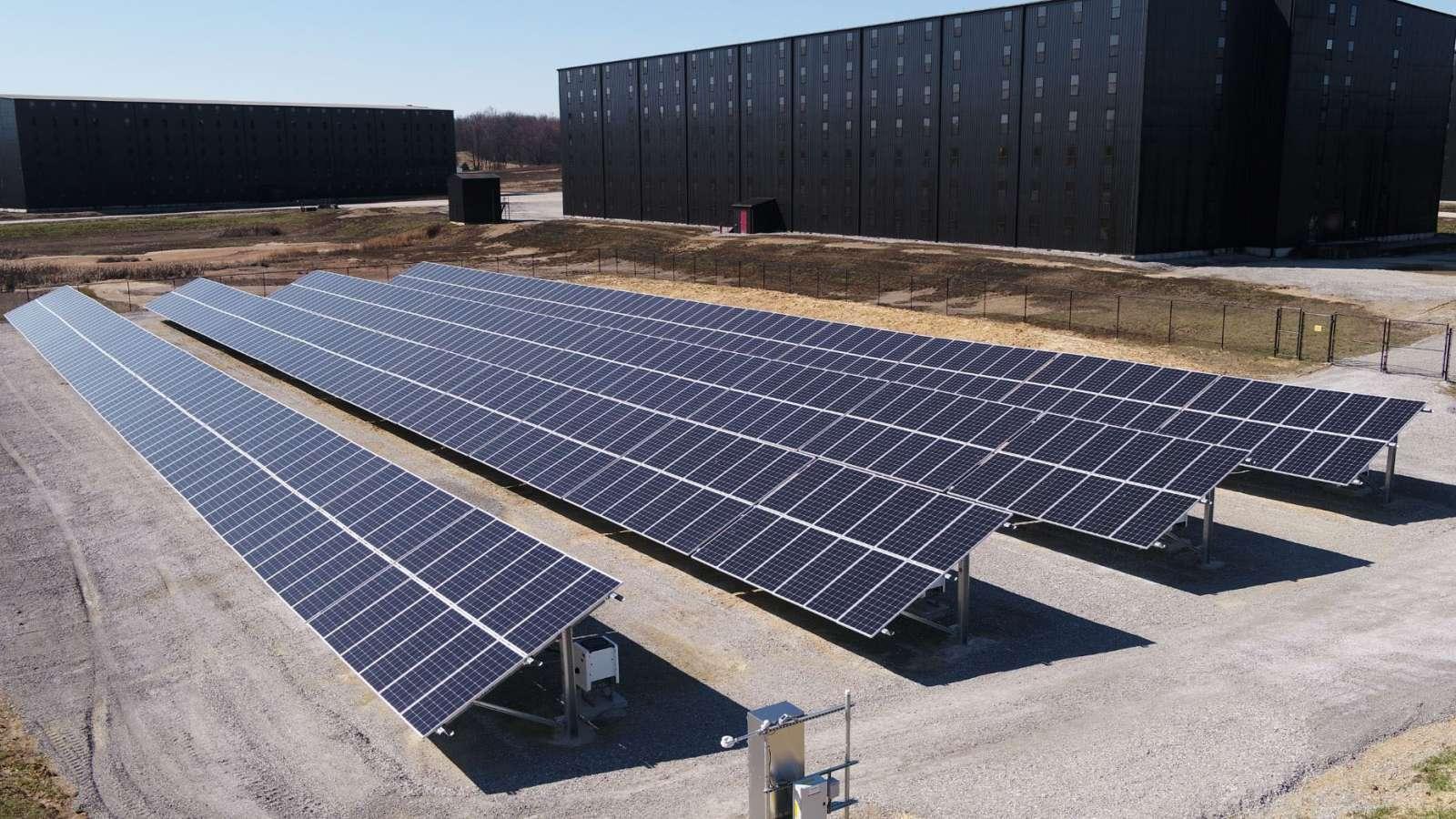 Rows of solar panels on a sunny day near large black buildings, surrounded by gravel and grass.