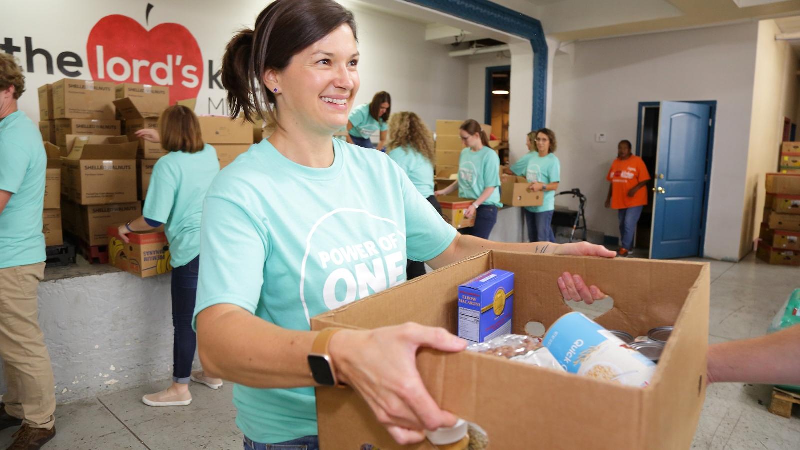 Smiling woman in a teal shirt holds a box of food items, helping with a group at a community food pantry.
