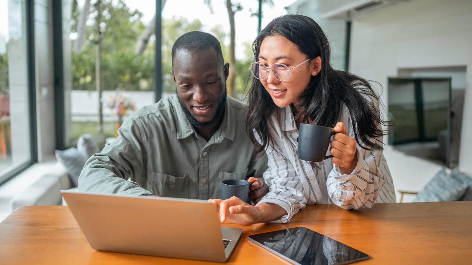 A man and woman smile while looking at a laptop. The woman holds a mug, and a tablet lies on the table in a bright, modern room.
