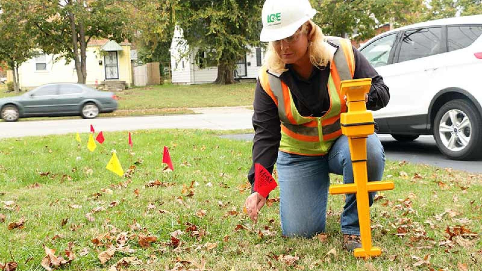 LG&E worker wearing a high-vis vest, safety goggles, and a hard hat placing red and yellow flag markers in a front yard.