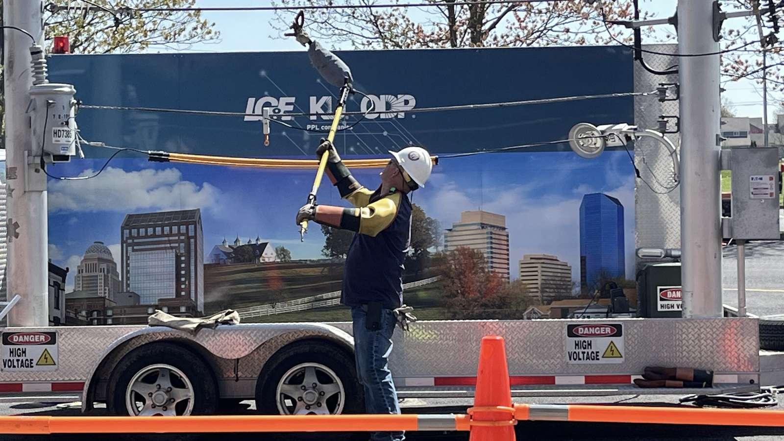Utility worker in protective gear using a tool on high voltage lines, set against backdrop on a trailer.