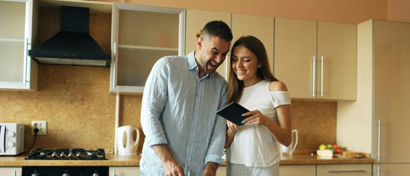 Smiling couple in a modern kitchen, chopping vegetables and looking at a tablet. Fresh fruits and vegetables are on the table.