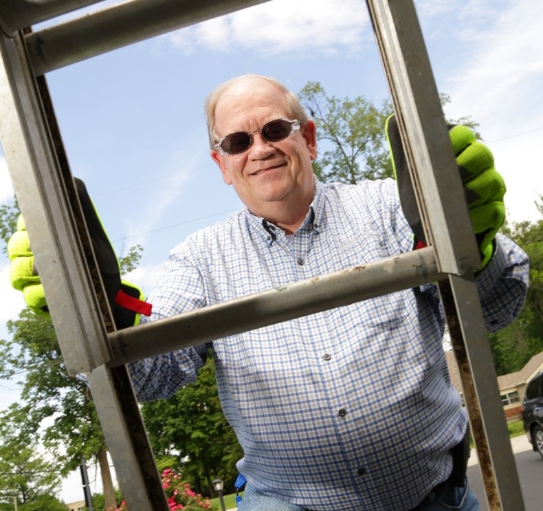 Smiling man wearing yellow gloves and sunglasses peers through the rungs of a ladder